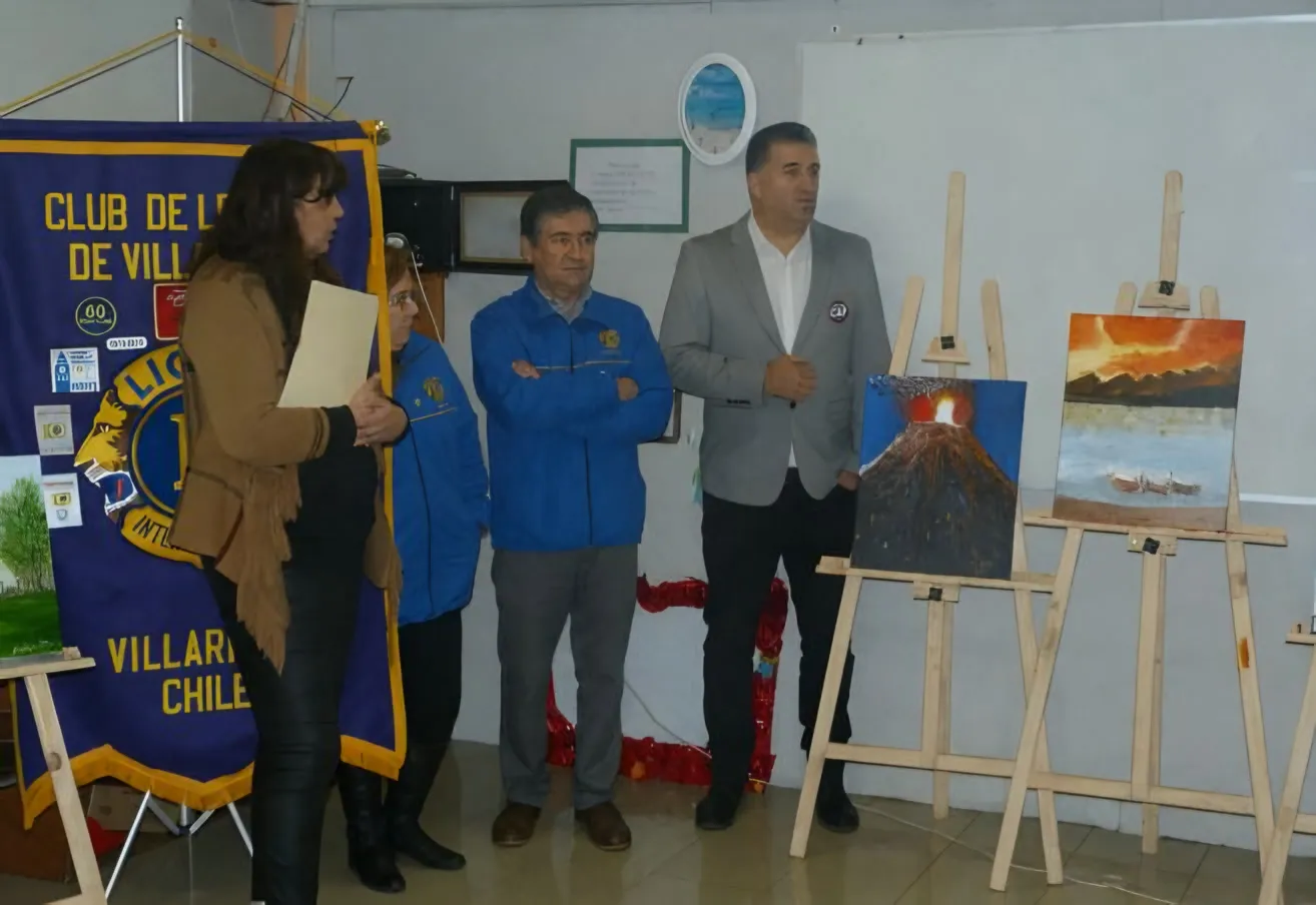 Organizadores junto a caballetes con pinturas al óleo y el estandarte del Club de Leones de Villarrica.