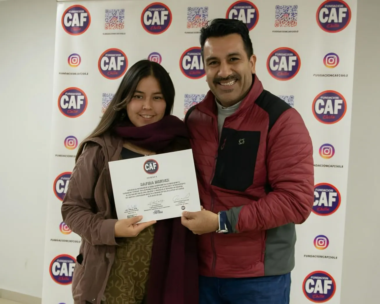 Dos personas adultas sostienen un diploma frente al telón de la fundación.